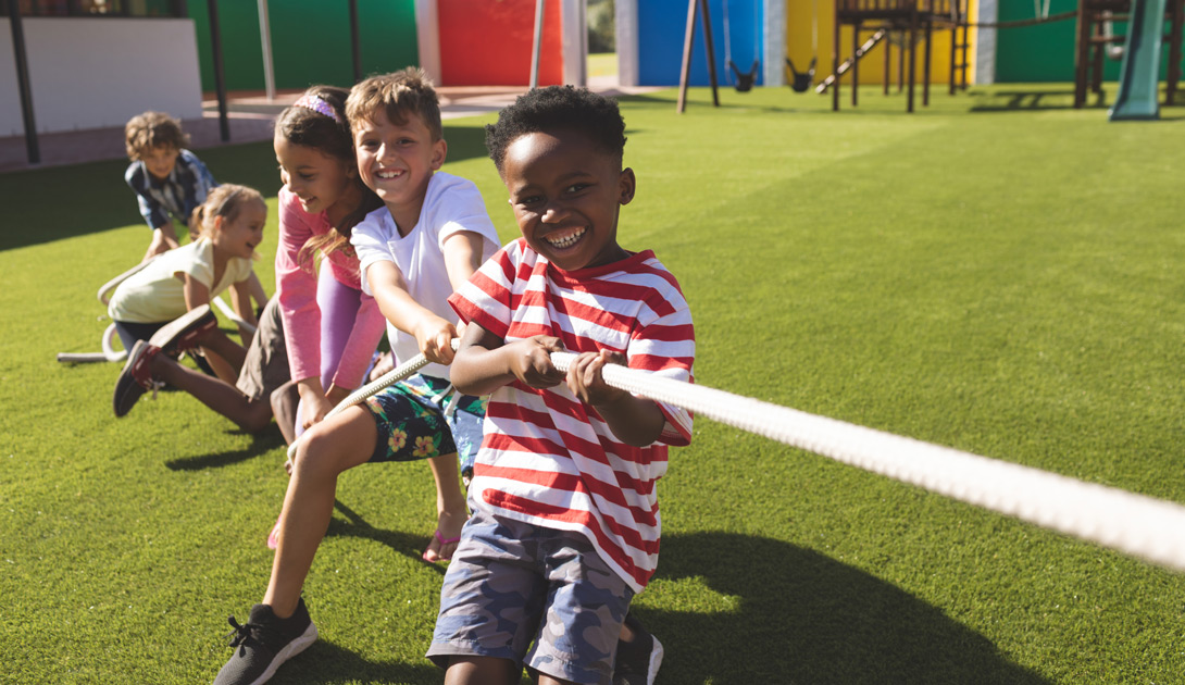 Front view of multi ethnic group of happy school kids playing tug of war in playground